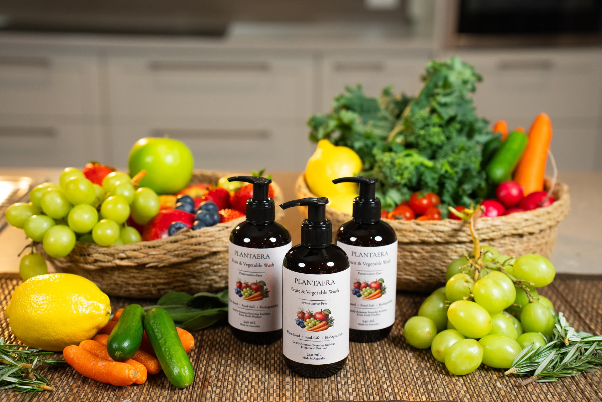 Three Plantaera bottles on a woven mat with baskets of fresh fruit and vegetables