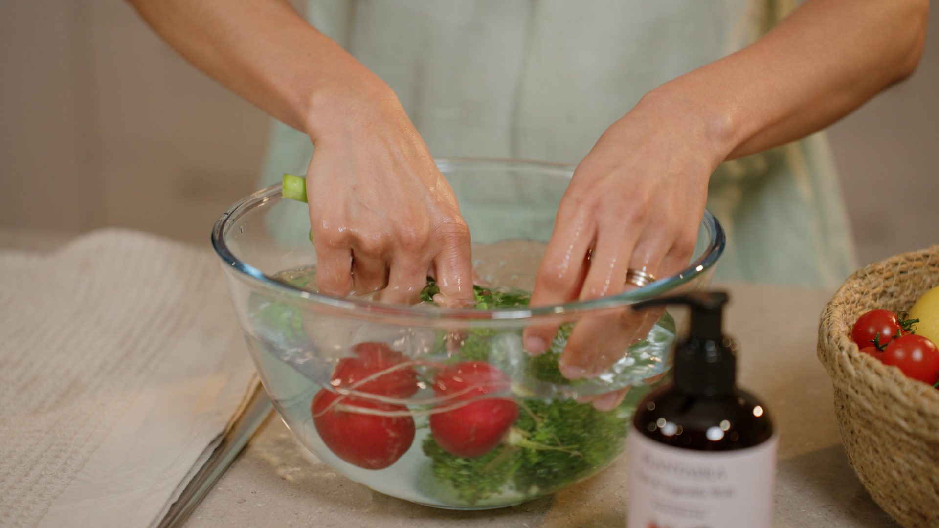 Hands rinsing radishes, broccoli and herbs in a glass bowl beside a Plantaera bottle