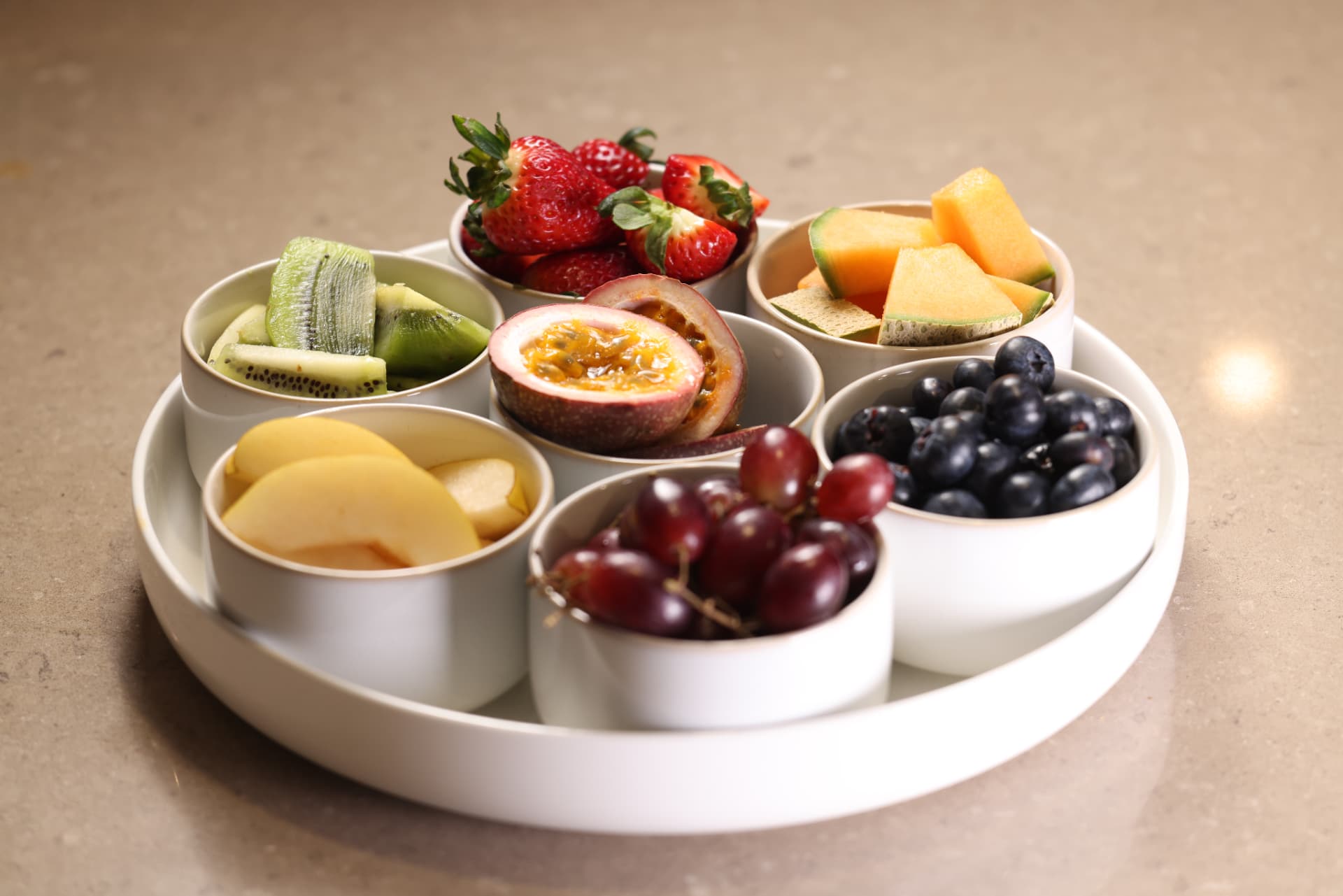 A round platter with bowls of fresh strawberries, melon, kiwi, grapes, blueberries and passionfruit