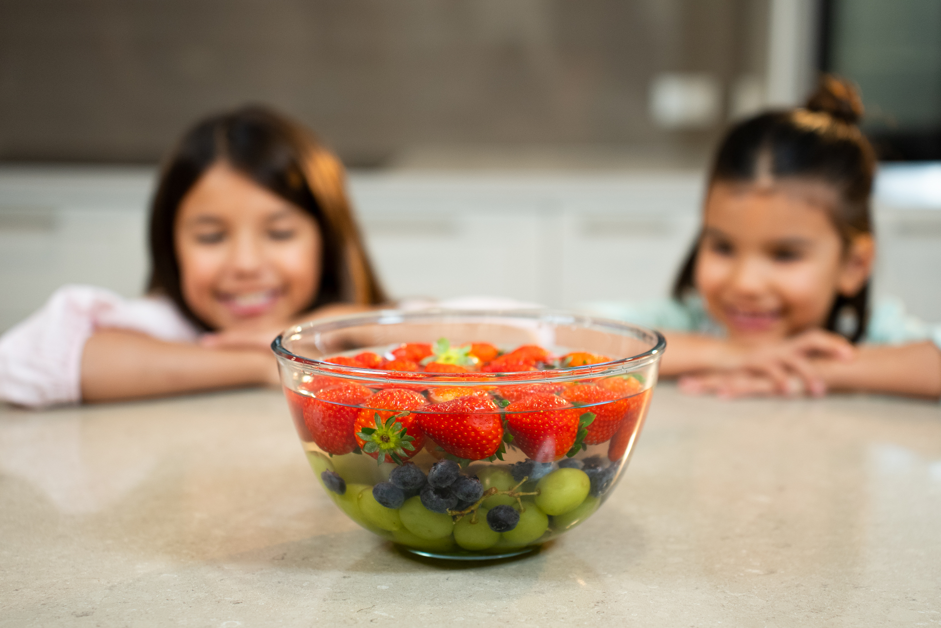 Two children smiling at a glass bowl of washed strawberries, grapes and blueberries